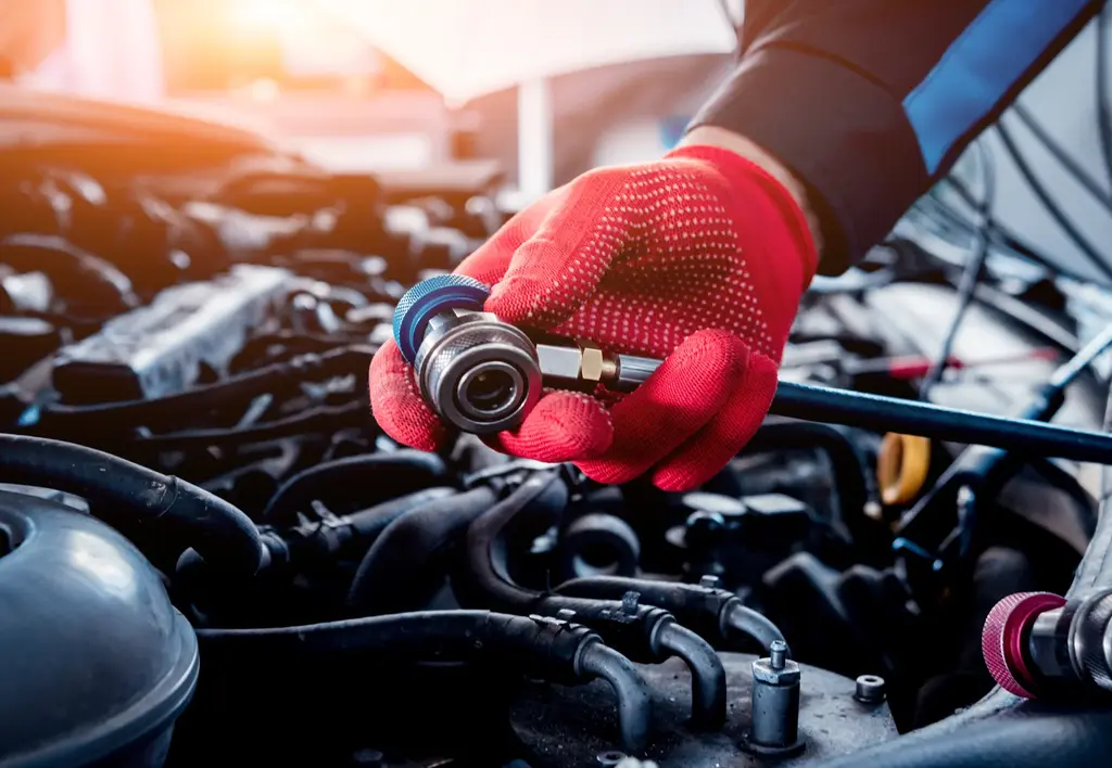 Main gantée de mécanicien tenant un raccord rapide de climatisation au dessus d'un moteur de voiture